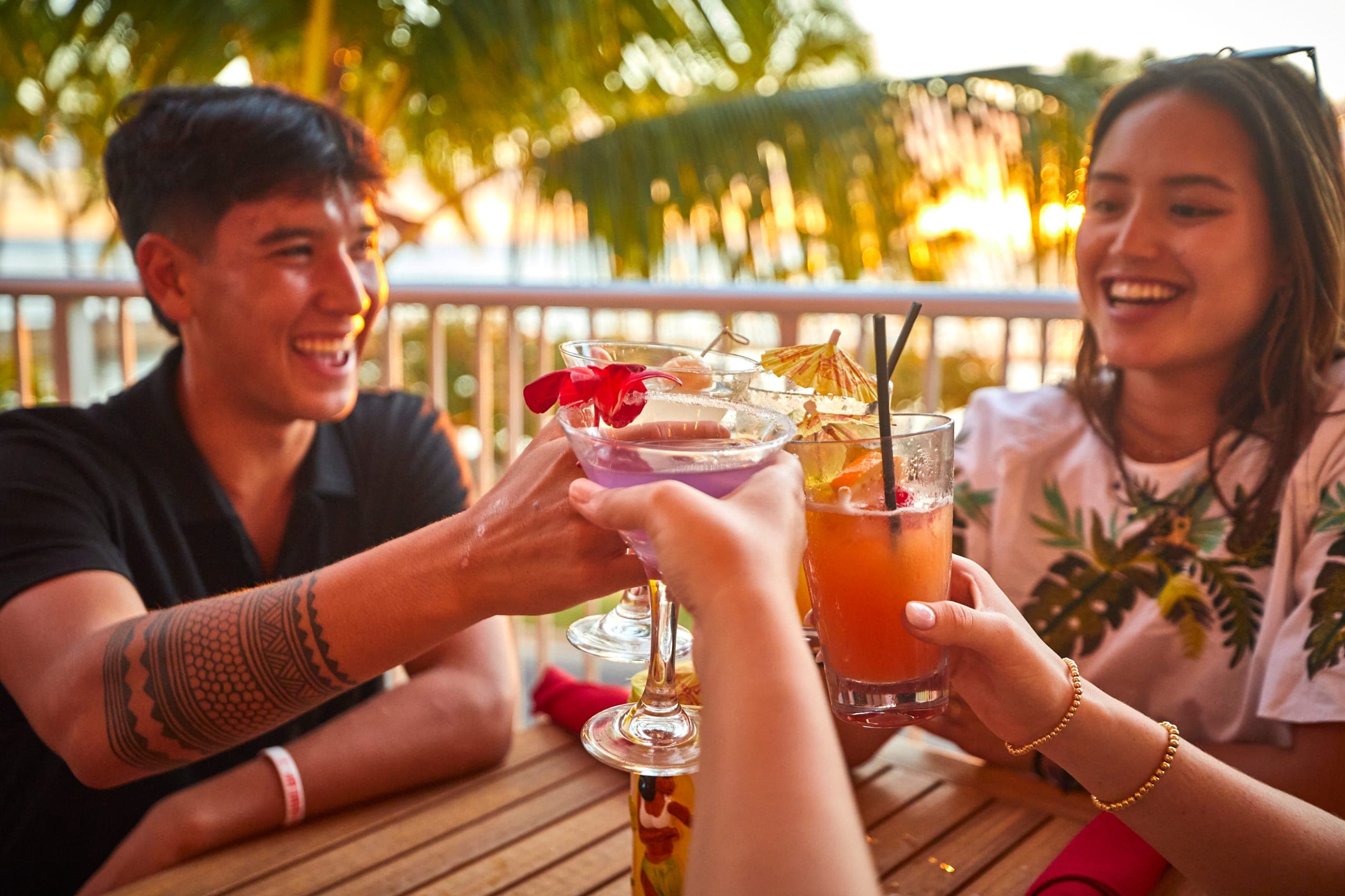 A young group of local friends toasting with a Butterfly Sunset Martini, Strawberry Squeeze, and Ube Colada at a beach-view rail table at Tiki's Grill & Bar.