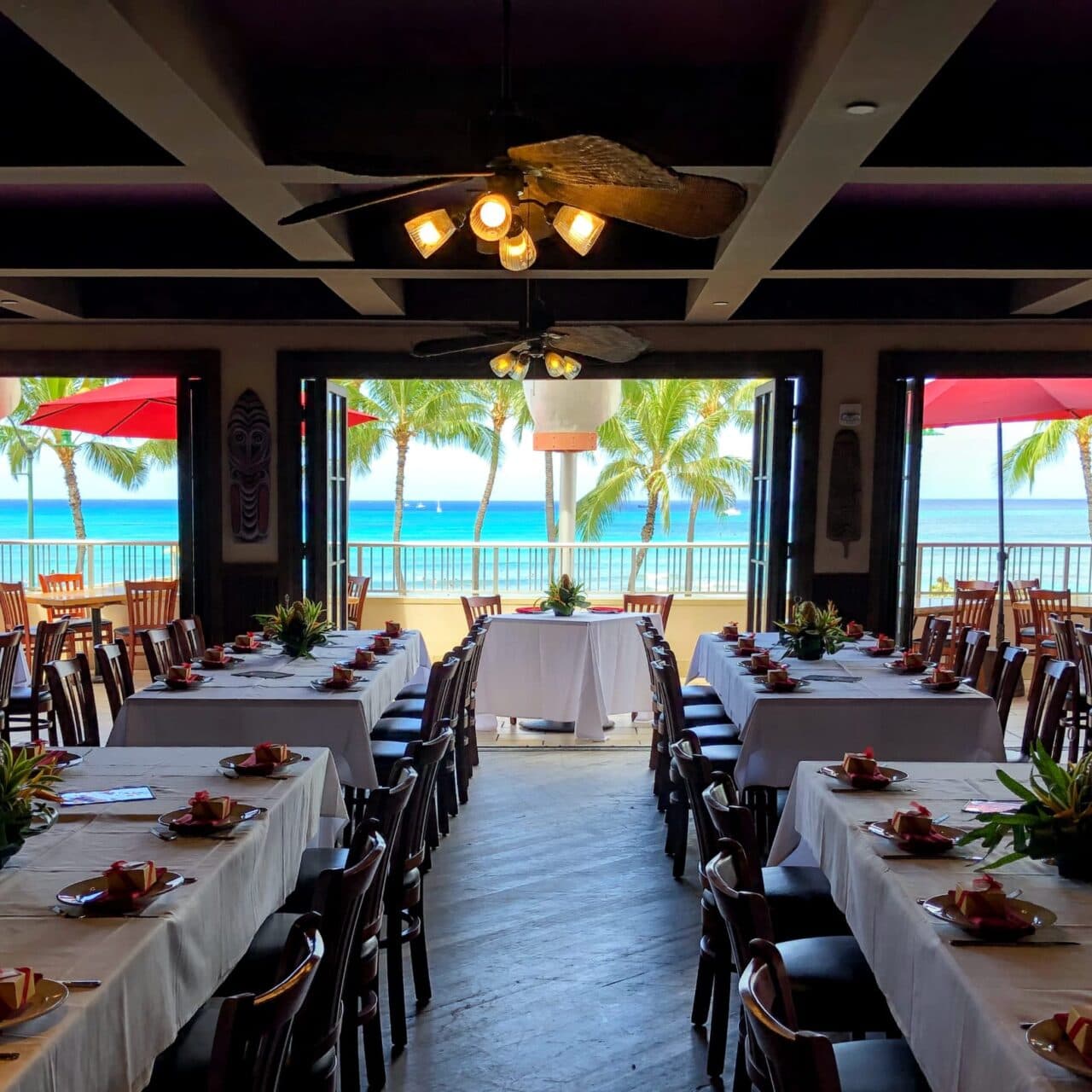 A large party table with white tablecloths and tropical flowers, set with an inside seating and an ocean view of the beach and coconut trees at Tiki's Grill & Bar.