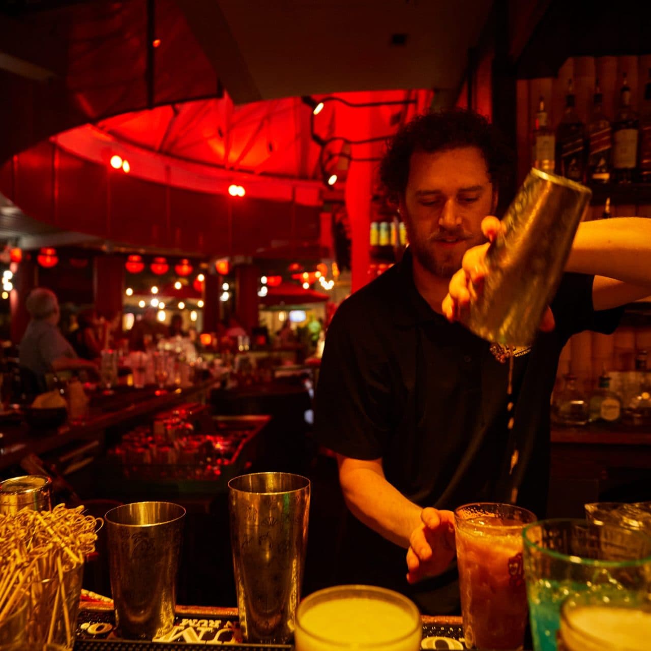 Bartender Austin at Tiki's Grill & Bar smiling behind the bar, straining a craft cocktail with a tray of fresh garnishes and house-made bar items.